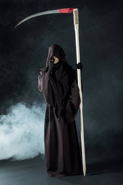 full length view of woman in death costume holding scythe and smoking cigarette on black