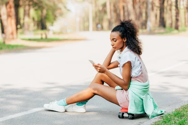 Menina Americana Africana Usando Smartphone Enquanto Sentado Skate Estrada — Fotografia de Stock