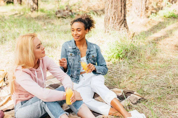 Two Multiethnic Girls Sitting Plaid Blanket Holding Bottles Orange Juice — Stock Photo, Image