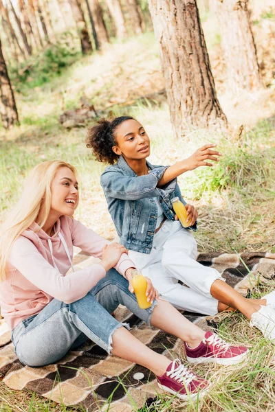 Dos Chicas Multiétnicas Sentadas Manta Cuadros Sosteniendo Botellas Jugo Naranja — Foto de Stock