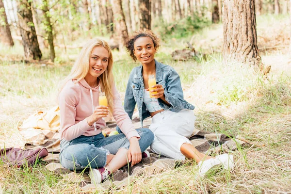 Two Multiethnic Girls Sitting Plaid Blanket Holding Bottles Orange Juice — Stock Photo, Image