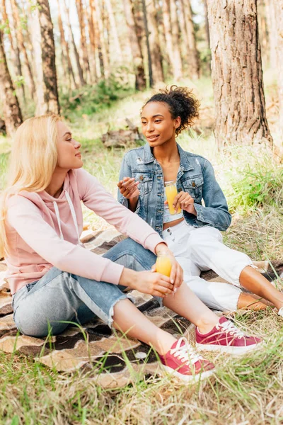 Duas Meninas Multiétnicas Sentadas Cobertor Xadrez Segurando Garrafas Suco Laranja — Fotografia de Stock