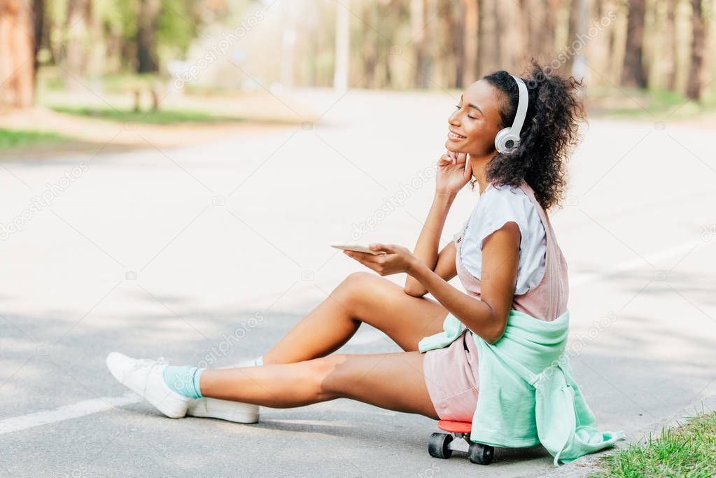 smiling african american girl listening music in headphones and using smartphone while sitting on penny board