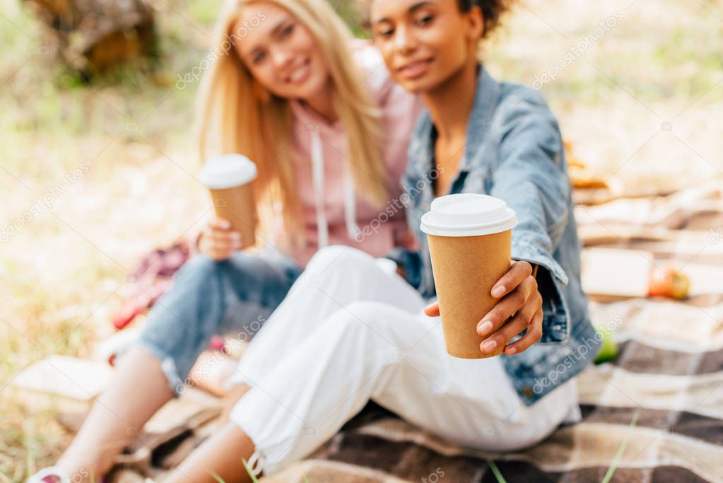 cropped view of two multiethnic friends sitting on plaid blanket and holding paper cups of coffee
