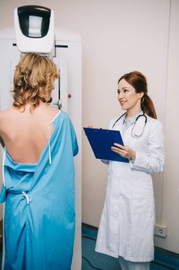 x-ray cihazında mamografi testi yapan hastanın yanında pano tutan gülümseyen radyograf
