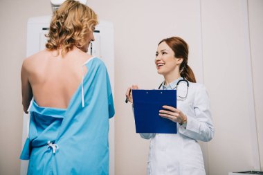 smiling radiologist holding clipboard while standing near patient making x-ray mammography test clipart