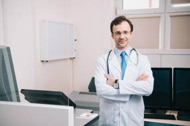 beyaz ceketli yakışıklı doktor çapraz kolları ile ayakta ve kameraya bakarak