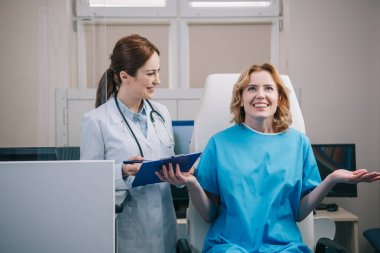 mutlu hasta panosu tutan gülümseyen doktor yakınında koltukta otururken gesturing