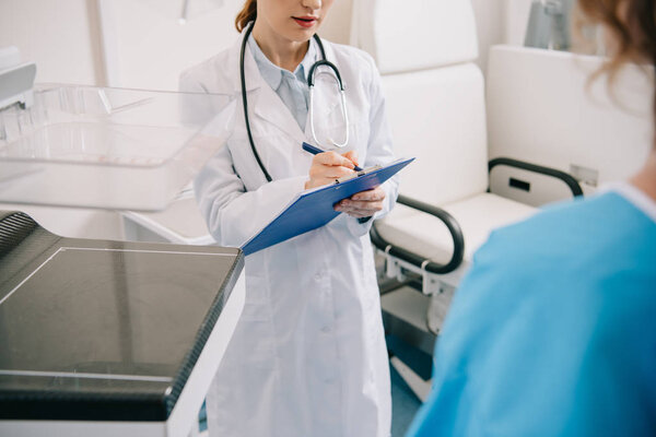 cropped view of doctor writing on clipboard while standing near patient 