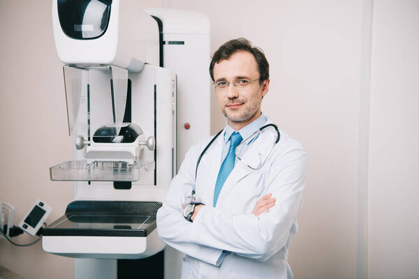 smiling doctor standing with crossed arms near x-ray machine and looking at camera