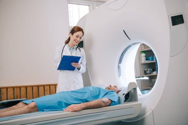 beautiful radiologist holding clipboard while preparing patient for tomography clipart