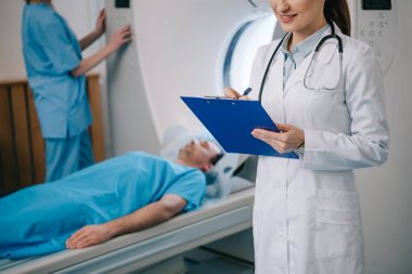cropped view of smiling radiographer writing on clipboard while assistant standing near lying patient and operating ct scanner clipart