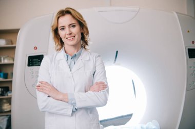 cheerful radiologist standing with crossed arms near computed tomography scanner and looking at camera clipart