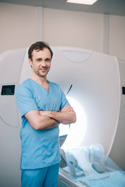 neşeli doktor çapraz kolları ile MRTarayıcı yanında duran ve kamera bakarak