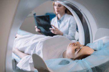 selective focus of radiologist holding x-ray diagnosis while patient lying on ct scanner bed during diagnostics clipart