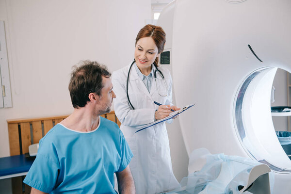 smiling radiologist writing on clipboard while standing near patient sitting on ct scanner bed