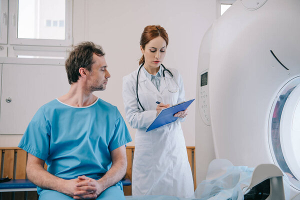 man sitting on ct scan bed near attentive radiologist writing on clipboard