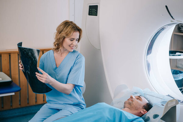 красивый улыбающийся доктор с радиологическим диагнозом и глядя на мужчину лежащего на кровати сканера КТ
