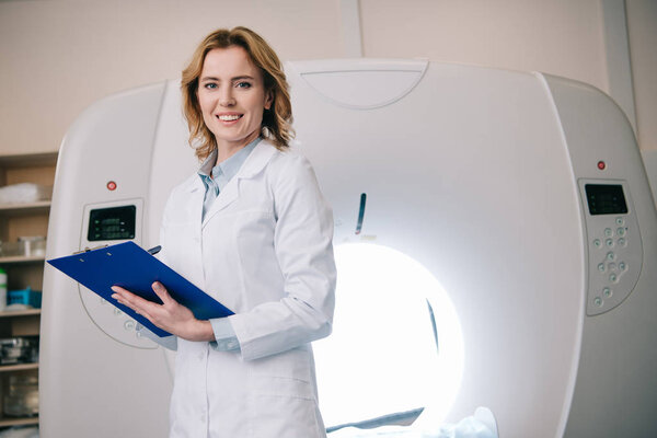 smiling radiologists writing on clipboard while standing near computed tomography scanner