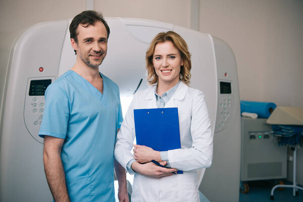улыбающимся врачам, смотрящим в камеру, стоя рядом со сканером компьютерной томографии
 