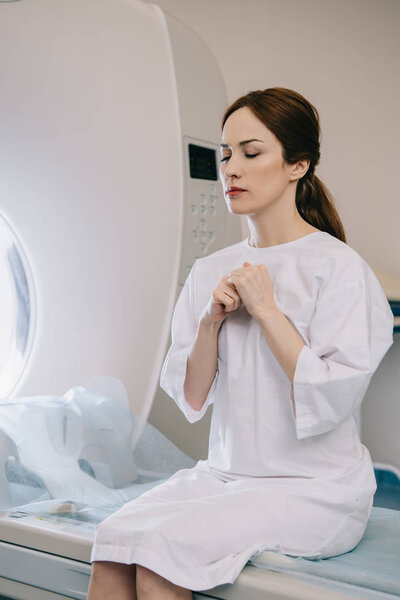 attractive woman praying with closed eyes while sitting on mri scanner bed in hospital