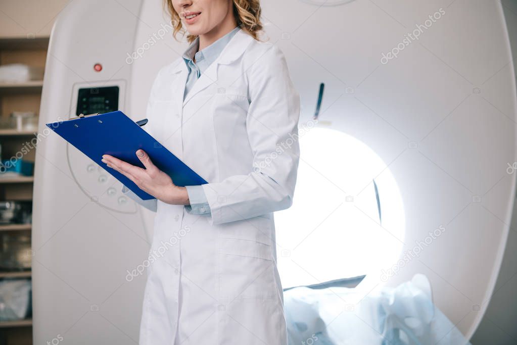 partial view of doctor writing on clipboard while standing near mri machine in hospital