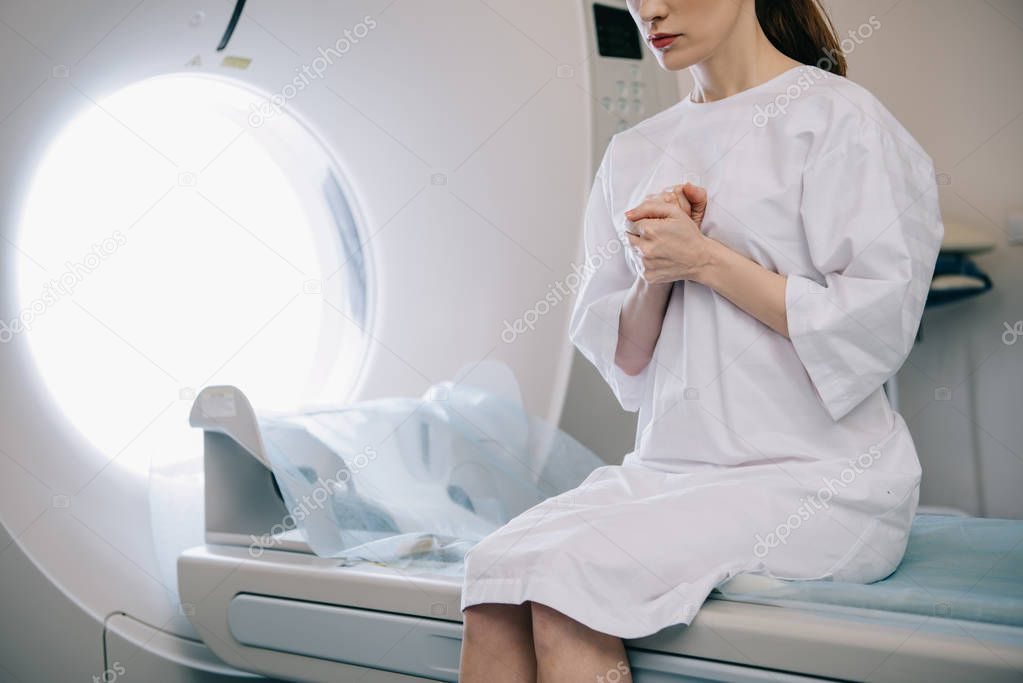 cropped view of woman showing pray gesture while sitting on computed tomography scanner bed