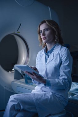 thoughtful radiologist holding digital tablet with x-ray diagnosis while sitting near computed tomography scanner and looking away clipart