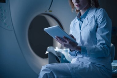 partial view of radiologist holding digital tablet with x-ray diagnosis while sitting near ct scanner clipart