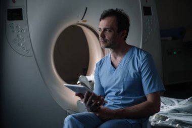 bilgisayarlı tomografi tarayıcı yakınında otururken x-ray tanısı ile dijital tablet tutan ciddi radyolog ve uzağa bakarak
