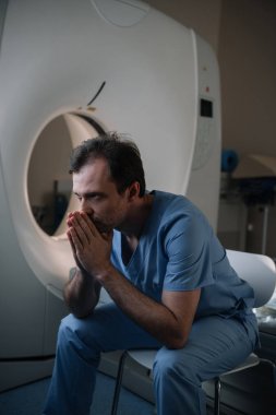 thoughtful radiologist sitting near computed tomography scanner in hospital and looking away clipart