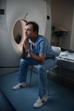serious doctor sitting near computed tomography scanner in hospital and looking up clipart