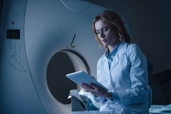 serious doctor looking at digital tablet with x-ray diagnosis while sitting near ct scanner 