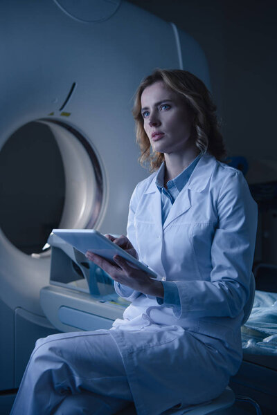 thoughtful radiologist holding digital tablet with x-ray diagnosis while sitting near computed tomography scanner and looking away