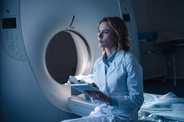 serious radiologist holding digital tablet while sitting near computed tomography scanner and looking away