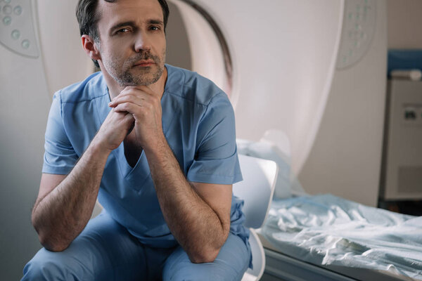 задумчивый врач сидит рядом с компьютерным томографом в больнице и смотрит в камеру
