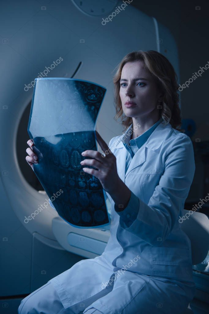 Serious doctor looking at x-ray diagnosis while sitting near ct scanner