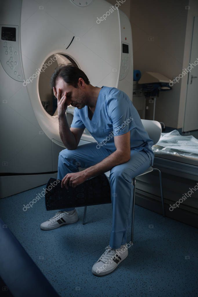 Exhausted radiologist holding x-ray diagnosis while sitting near ct scanner in hospital