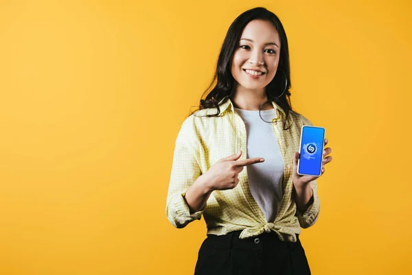乌克兰 2019年4月16日 迷人的微笑的亚洲女孩指着智能手机与Shazam应用程序 隔离在黄色 — 图库照片