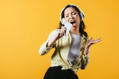 attractive asian girl singing and listening music with headphones and smartphone, isolated on yellow