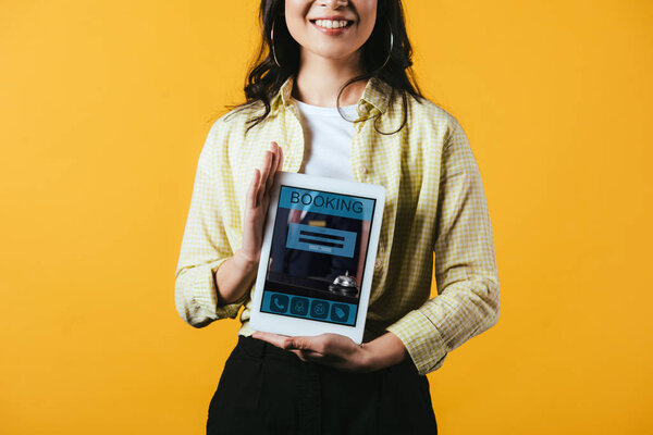 обрезанный вид улыбающейся девушки, показывающей цифровой планшет с приложением бронирования, изолированный на желтый
