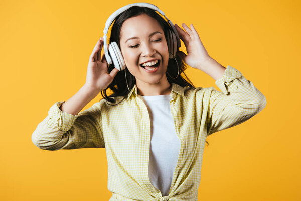 attractive asian girl singing and listening music with headphones, isolated on yellow