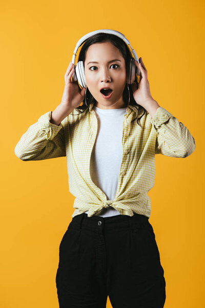 shocked asian girl listening music with headphones, isolated on yellow