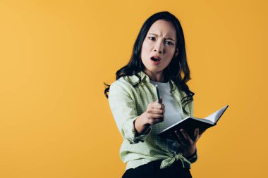 angry asian woman with notebook and pen, isolated on yellow  clipart