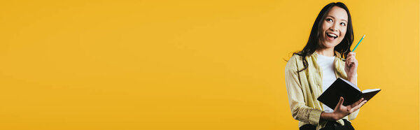 улыбающийся азиатский студент с ноутбуком и ручкой, изолированный на желтом
 