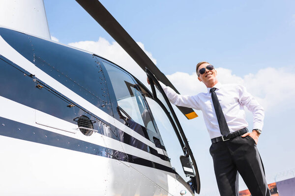 handsome Pilot in formal wear and sunglasses smiling near helicopter