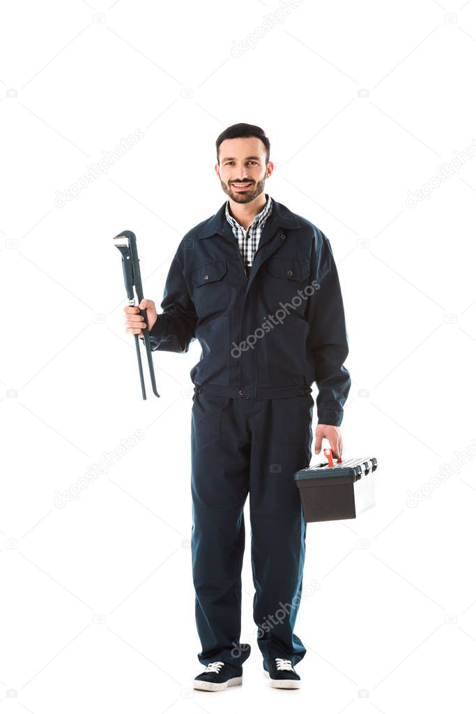 happy plumber in overalls holding toolbox and adjustable wrench while smiling at camera isolated on white