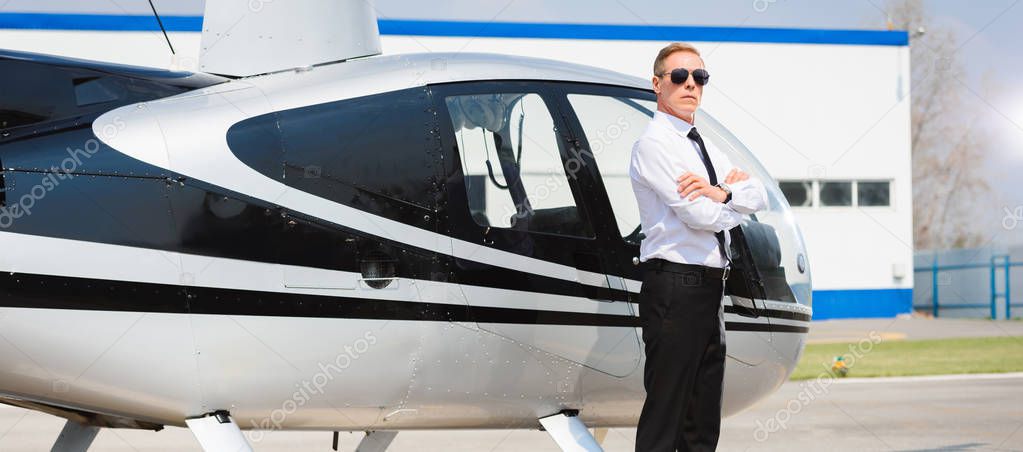 panoramic shot of Pilot in sunglasses and formal wear with crossed arms near helicopter