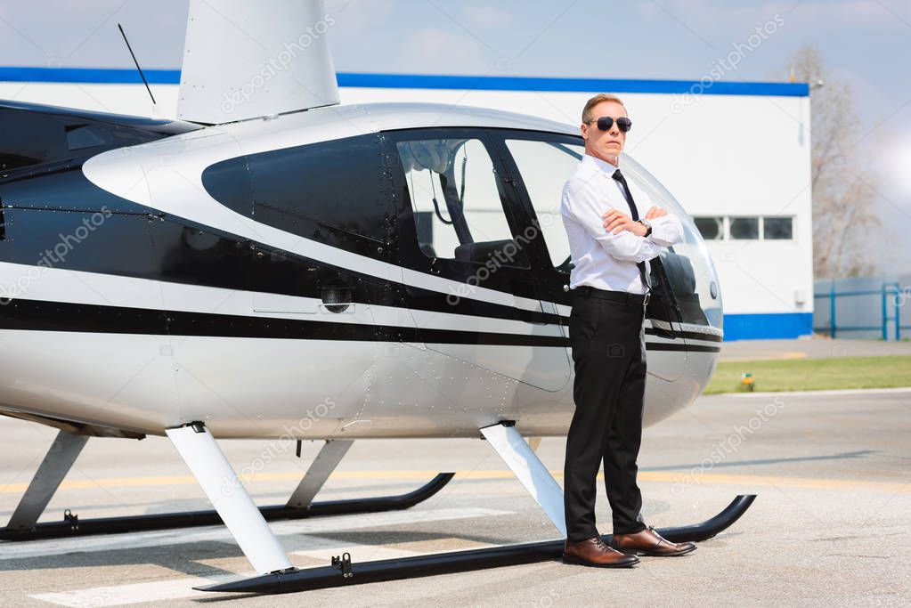 Pilot in sunglasses and formal wear with crossed arms posing near helicopter