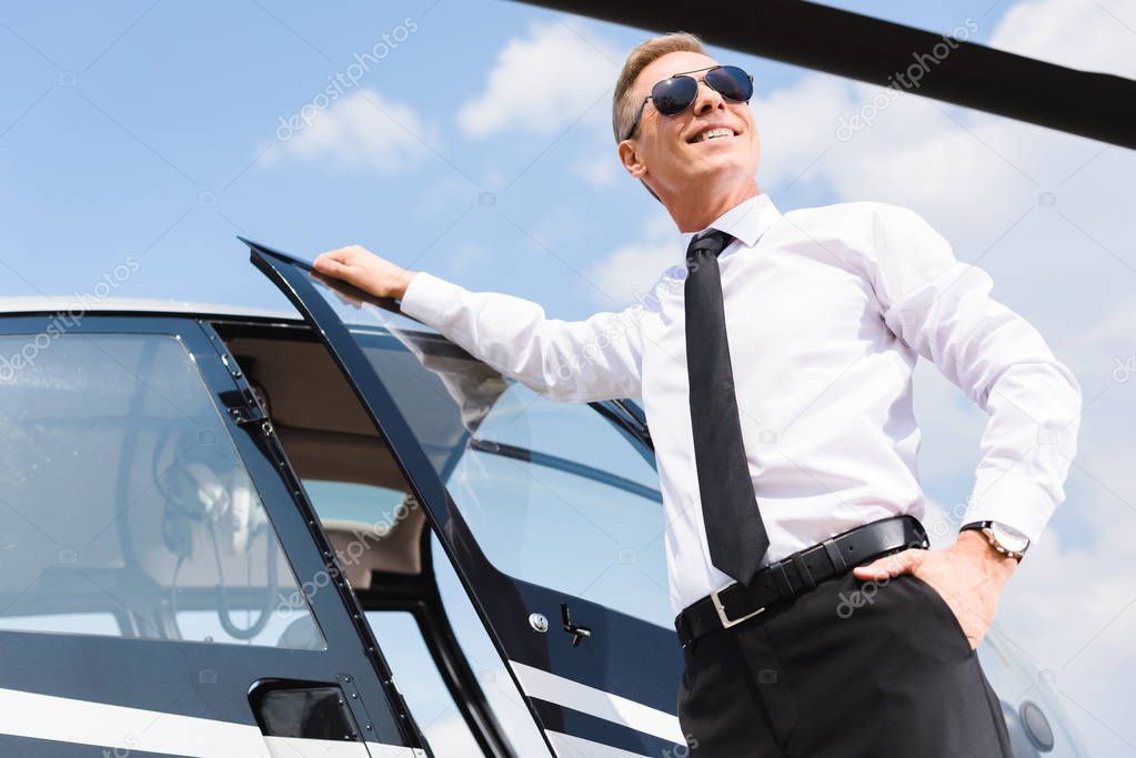 low angle view of handsome smiling Pilot in formal wear and sunglasses near helicopter